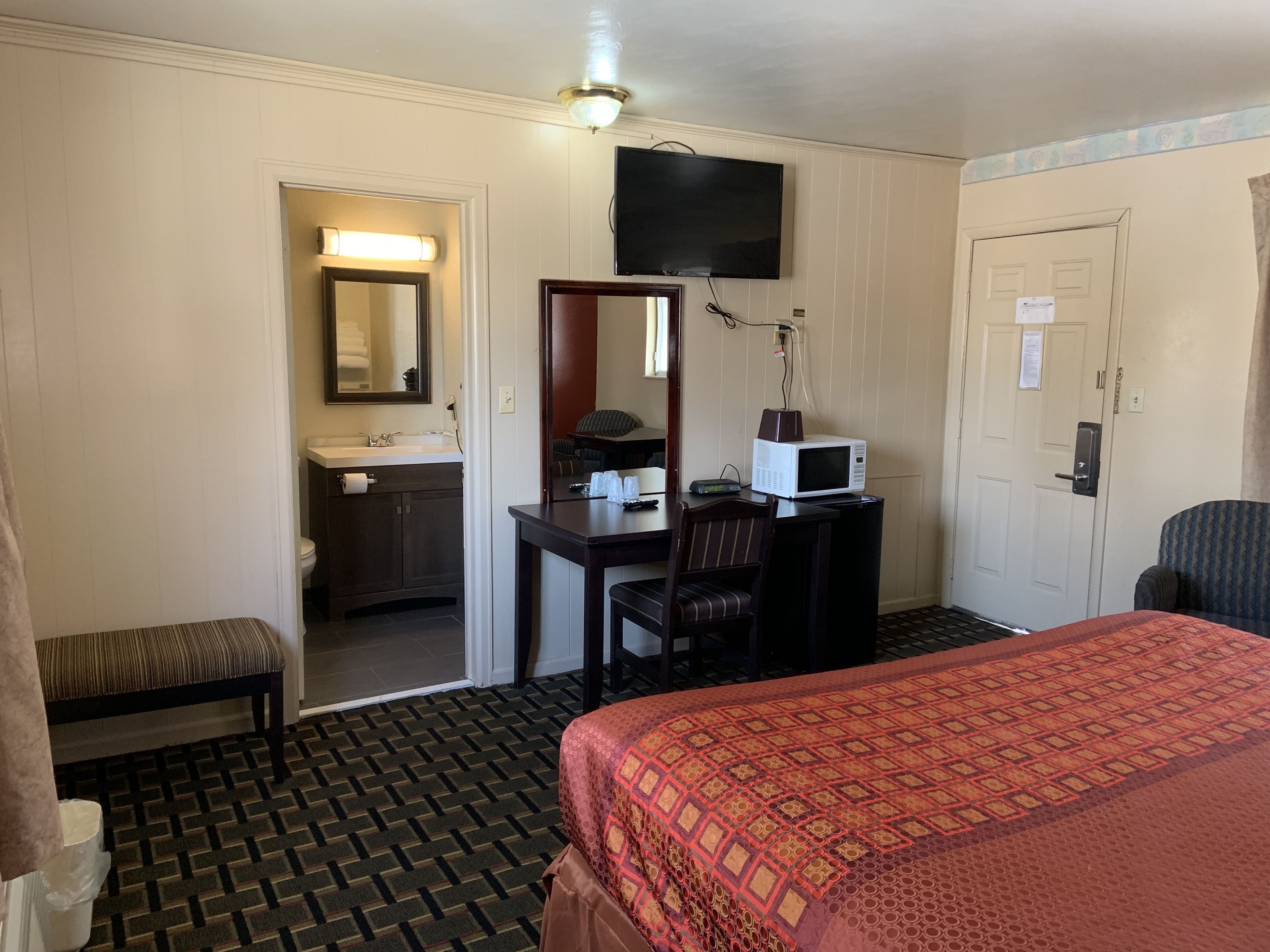 A hotel room with a bed, a desk with a chair, a television, a microwave, a bathroom with a sink and mirror, and a patterned carpet.
