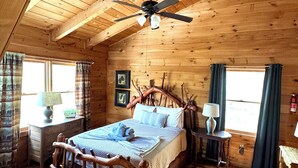 Ein Schlafzimmer einer Holzhütte mit einem Bett, einem Deckenventilator, einem Nachttisch, einer Lampe und einem Fenster mit Vorhängen.