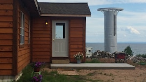 Une cabane en bois avec une porte blanche, un petit porche agrémenté de plantes en pot et, au fond, un grand château d’eau.