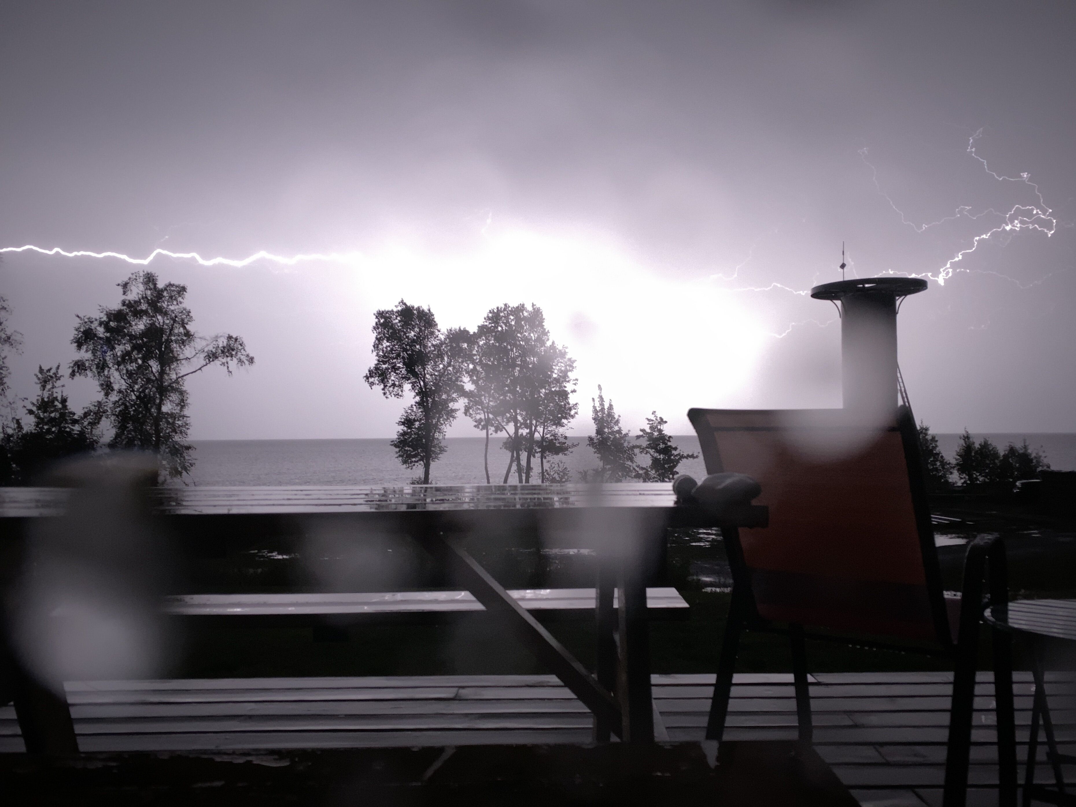Une terrasse avec une table et des chaises donnant sur une étendue d’eau, et où l’on peut voir des éclairs dans le ciel.