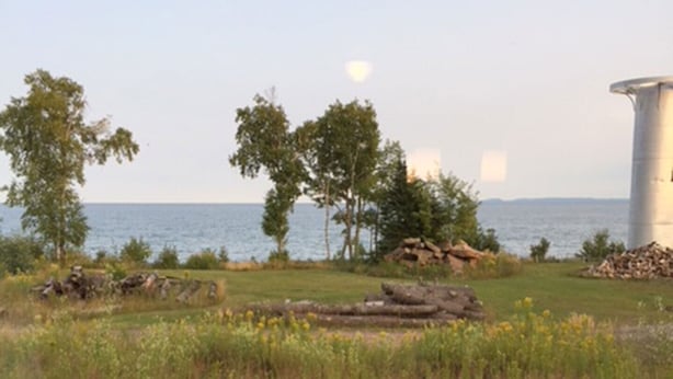 Vue depuis une pièce où l’on aperçoit une chaise en bois et une table, donnant sur un espace verdoyant avec des arbres et une étendue d’eau.