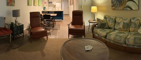 A living room with a sofa, two chairs, a coffee table, a TV, and a kitchen area in the background.