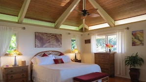 A bedroom with a wooden ceiling, a bed with white bedding, two bedside tables with lamps, a wooden bench, a dresser, and a potted plant.
