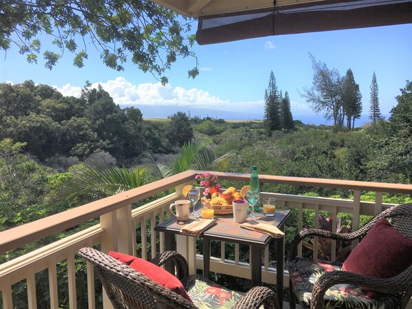 A balcony with wicker chairs, a table with breakfast items, and a view of a forest and ocean.