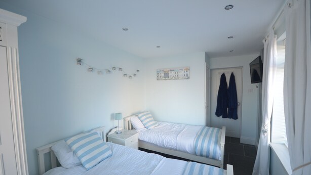 A bedroom with two beds, a nightstand, a wall-mounted TV, and a door with a coat rack. - SEASIDE COTTAGE WITH SEA VIEWS (Newbiggin-by-the-Sea)