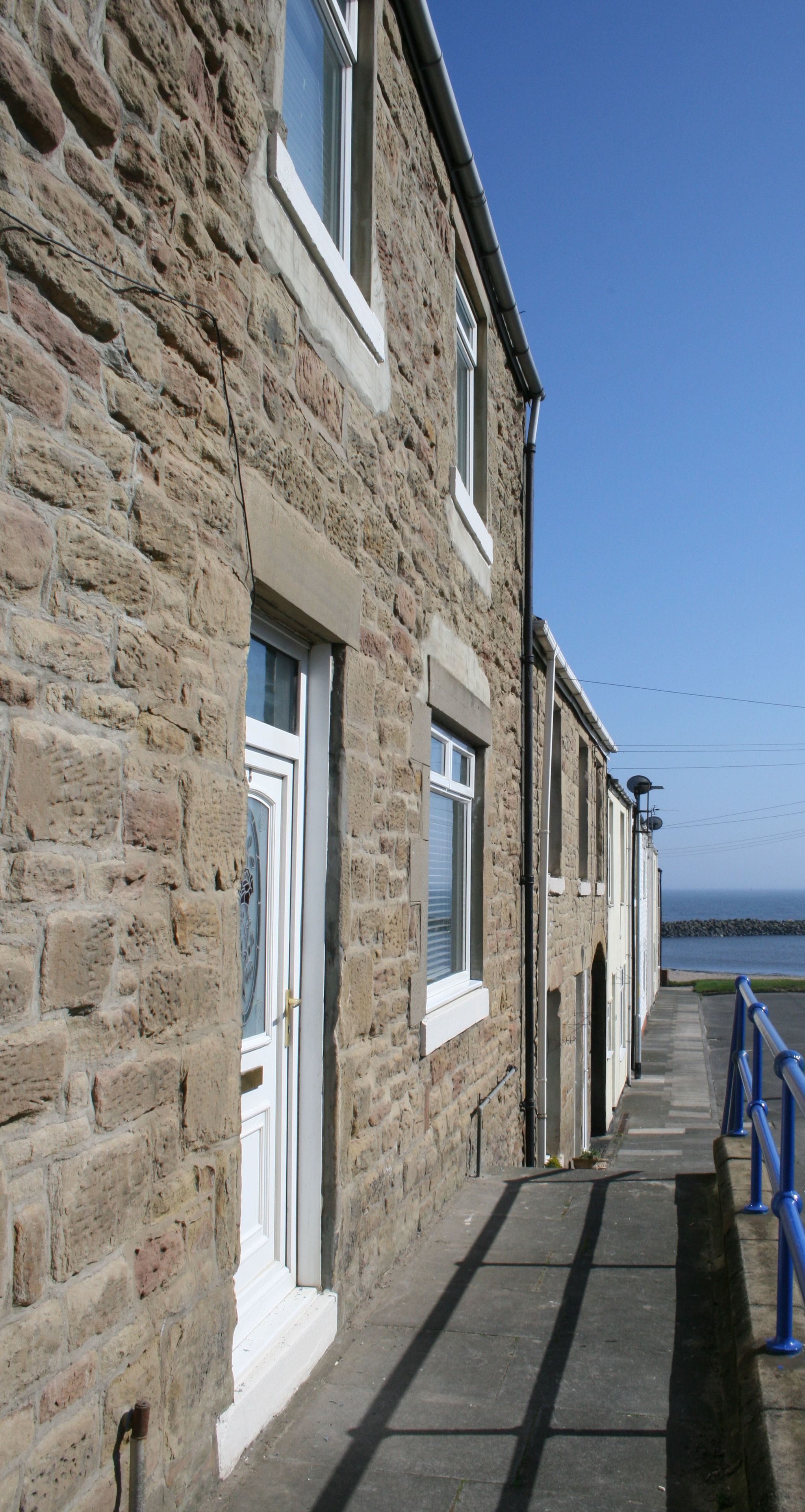 A stone building with white-framed windows, a blue railing, and a view of the sea.
