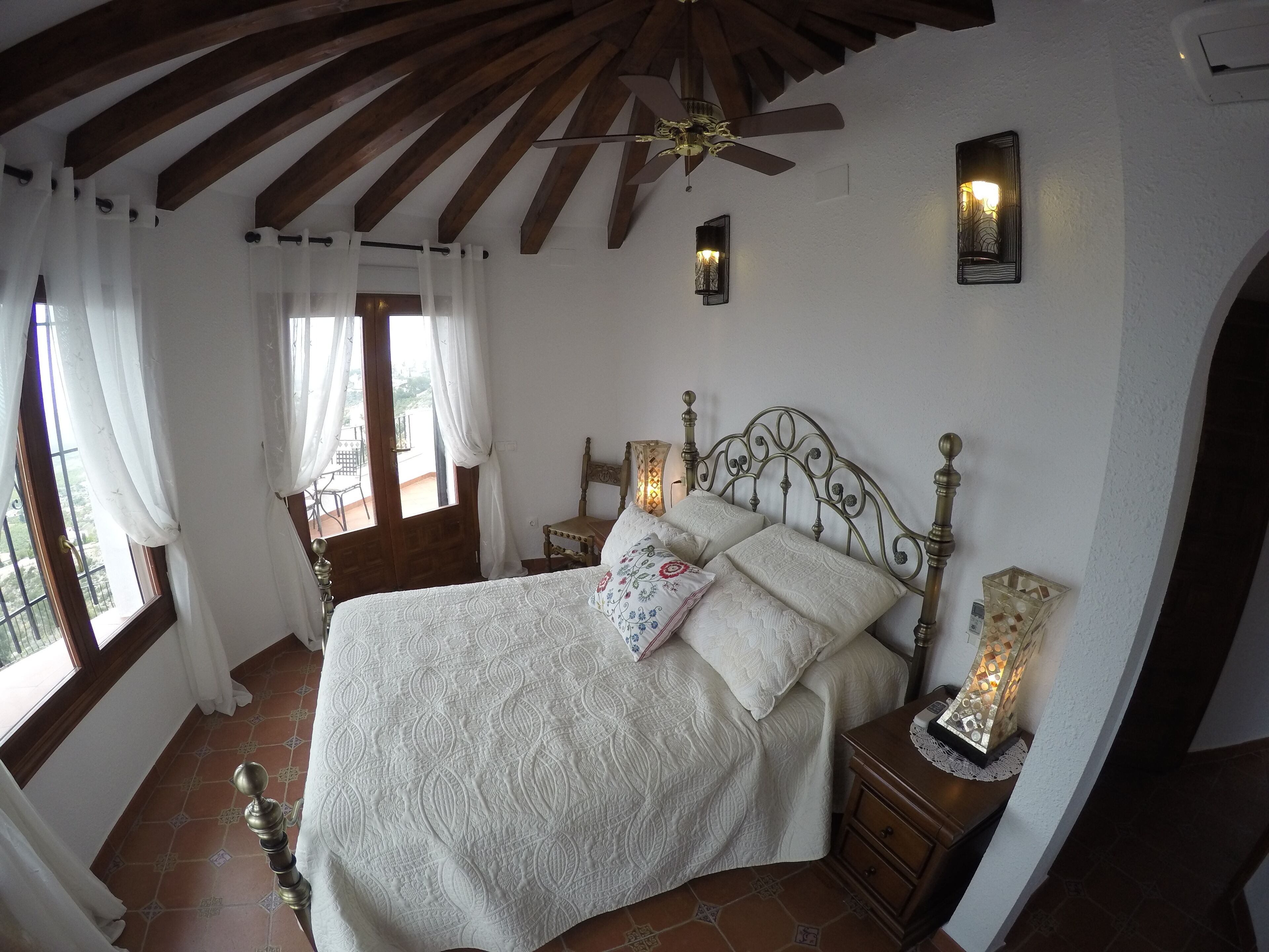 Un dormitorio con una cama, dos mesitas de noche, un velador, un ventilador de techo y una ventana con cortinas.