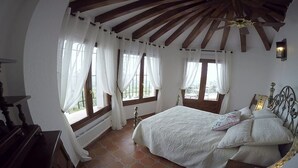 Un dormitorio con techo de madera, una cama con colcha estampada, dos ventanas con cortinas blancas y vistas al exterior.