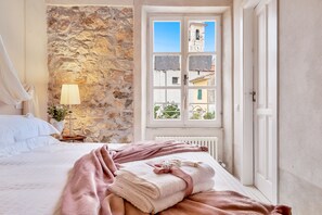 Une chambre avec un mur en pierre, une fenêtre donnant sur des immeubles, un lit avec du linge de lit blanc et une lampe de chevet.