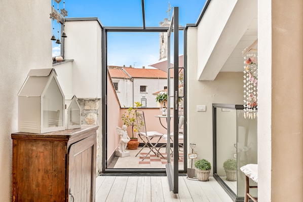 Un étroit couloir extérieur avec une armoire en bois, une porte vitrée donnant sur un patio aménagé avec une table, des chaises et des plantes en pot.