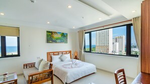 A hotel room with a large bed, a wooden chair, a small table, and a view of tall buildings through the window. - Salamander Hotel and Apartment (Da Nang)