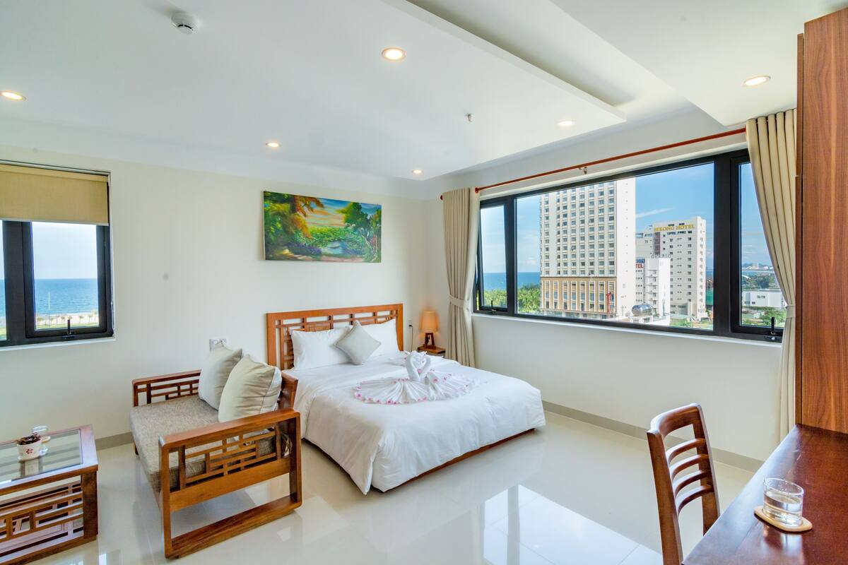 Quarto de hotel com uma cama grande, uma cadeira de madeira, uma mesa pequena e vista para edifícios altos pela janela.