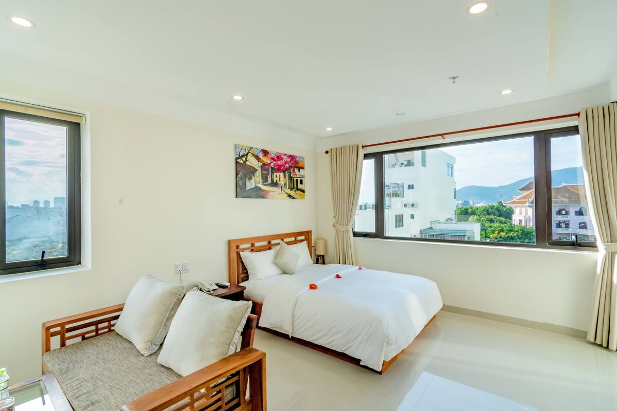 Quarto de hotel com cama, sofá, um quadro na parede e amplas janelas com cortinas.