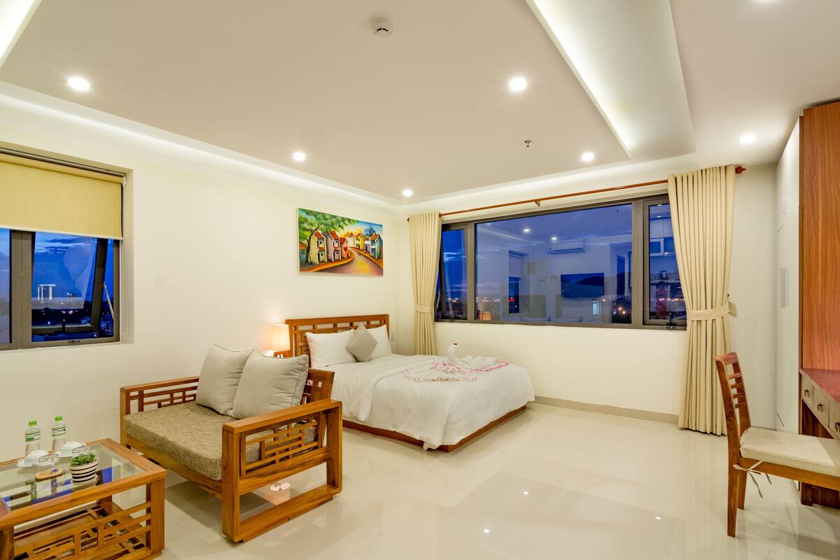 Quarto de hotel moderno com cama, sofá, cadeira de madeira, mesa de vidro e um quadro na parede.