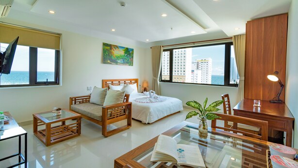 A modern hotel room with a bed, a sofa, a dining table, and a desk. There is a window with a view of the city and a plant on the table. - Salamander Hotel and Apartment (Da Nang)