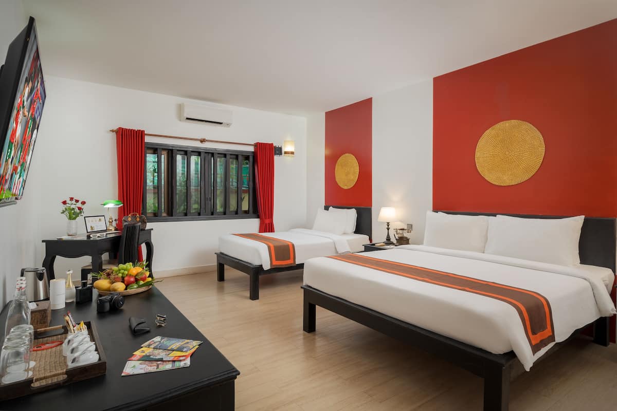 A hotel room with two beds, a desk with a fruit basket, a TV, and a window with red curtains.