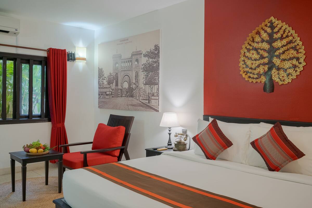 A hotel room with a bed, a chair, a table with fruit, a lamp, and a wall with a tree and a picture.