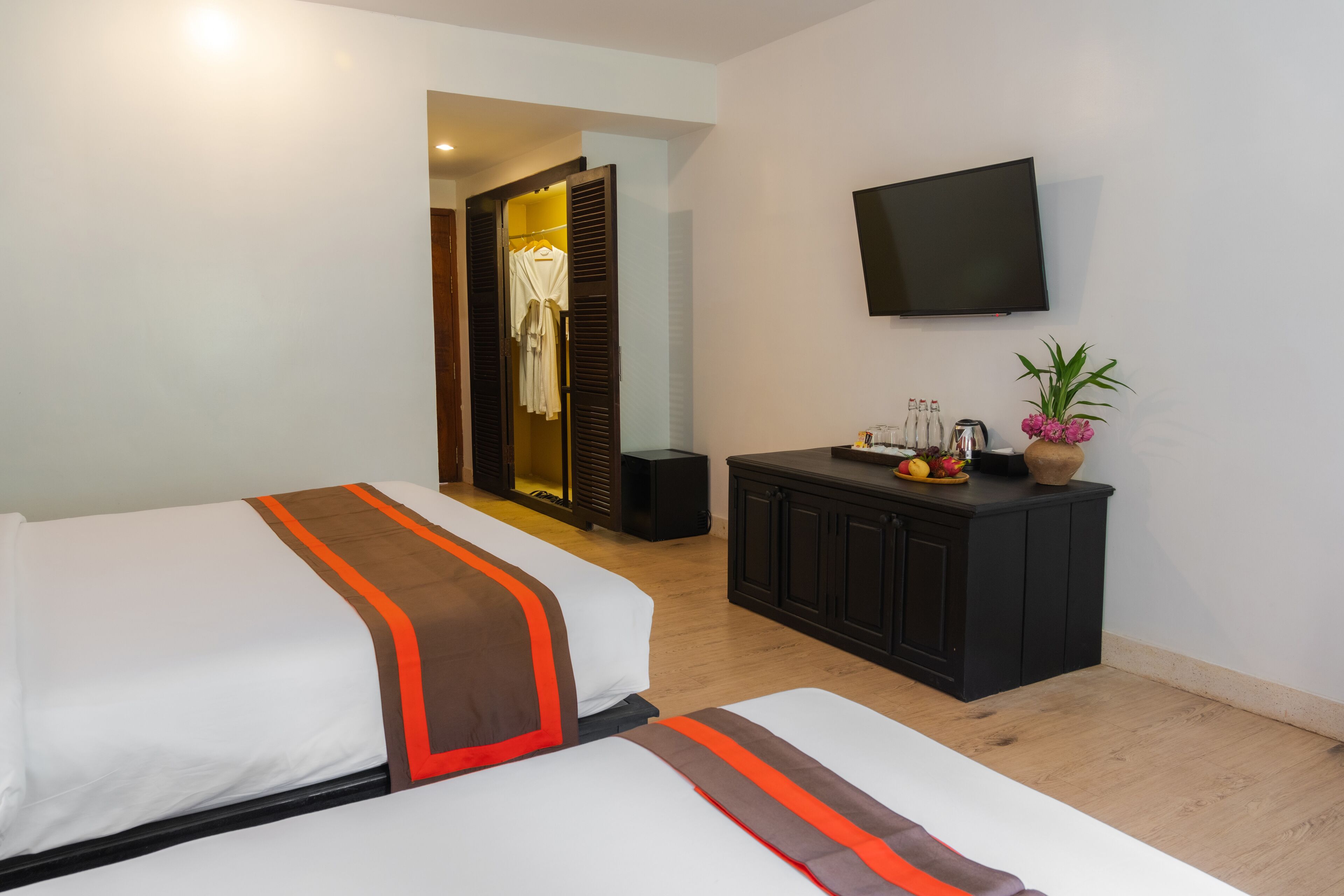 A hotel room with two beds, a TV, a wardrobe, and a small cabinet with fruit and flowers.