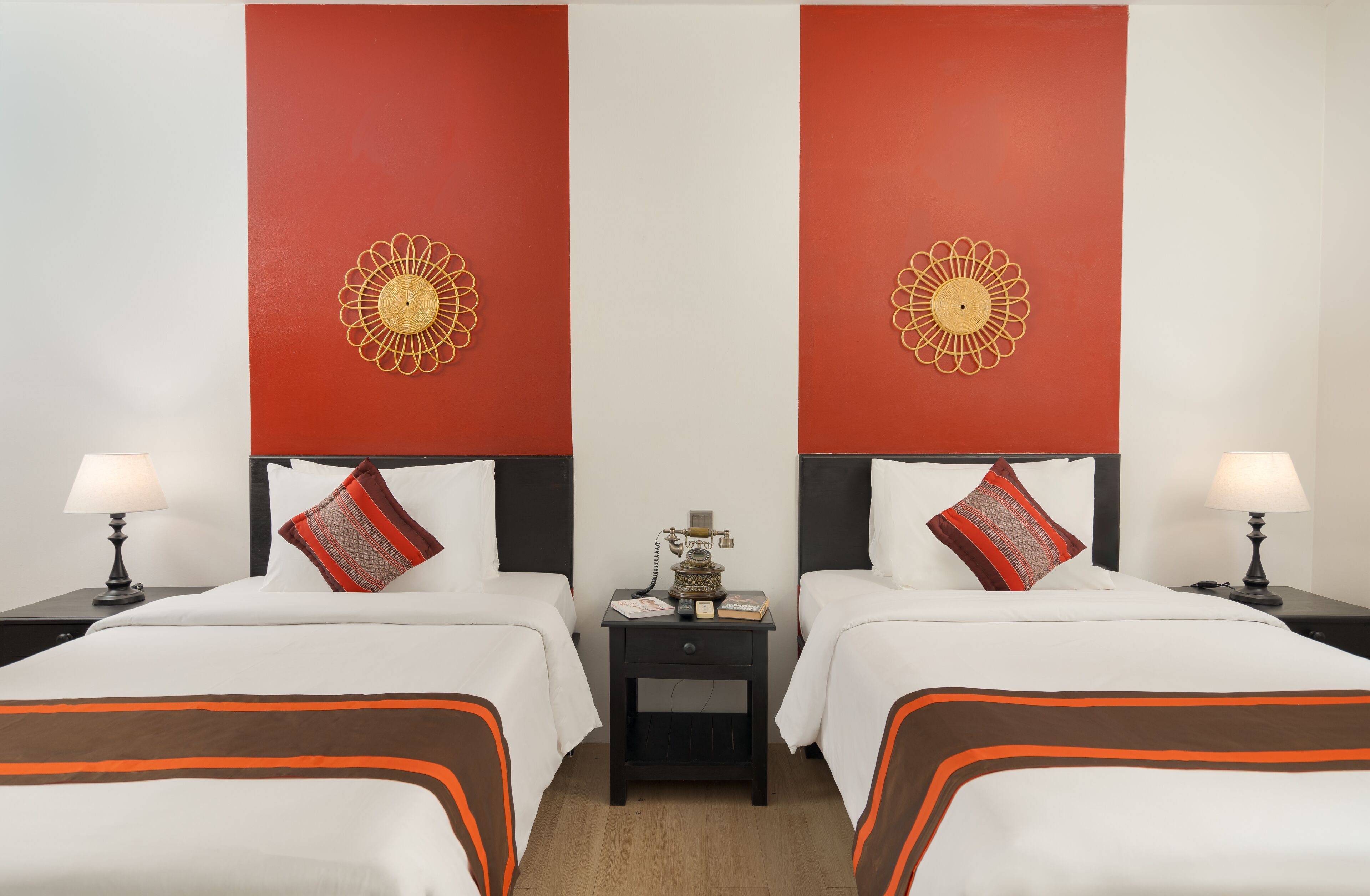 A hotel room with two beds, each with white bedding and orange accents, a black nightstand with a lamp, and a decorative wall piece above each bed.