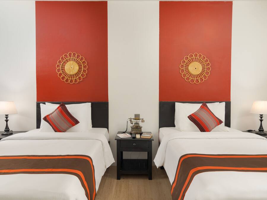 A hotel room with two beds, each with white bedding and orange accents, a black nightstand with a lamp, and a decorative wall piece above each bed.