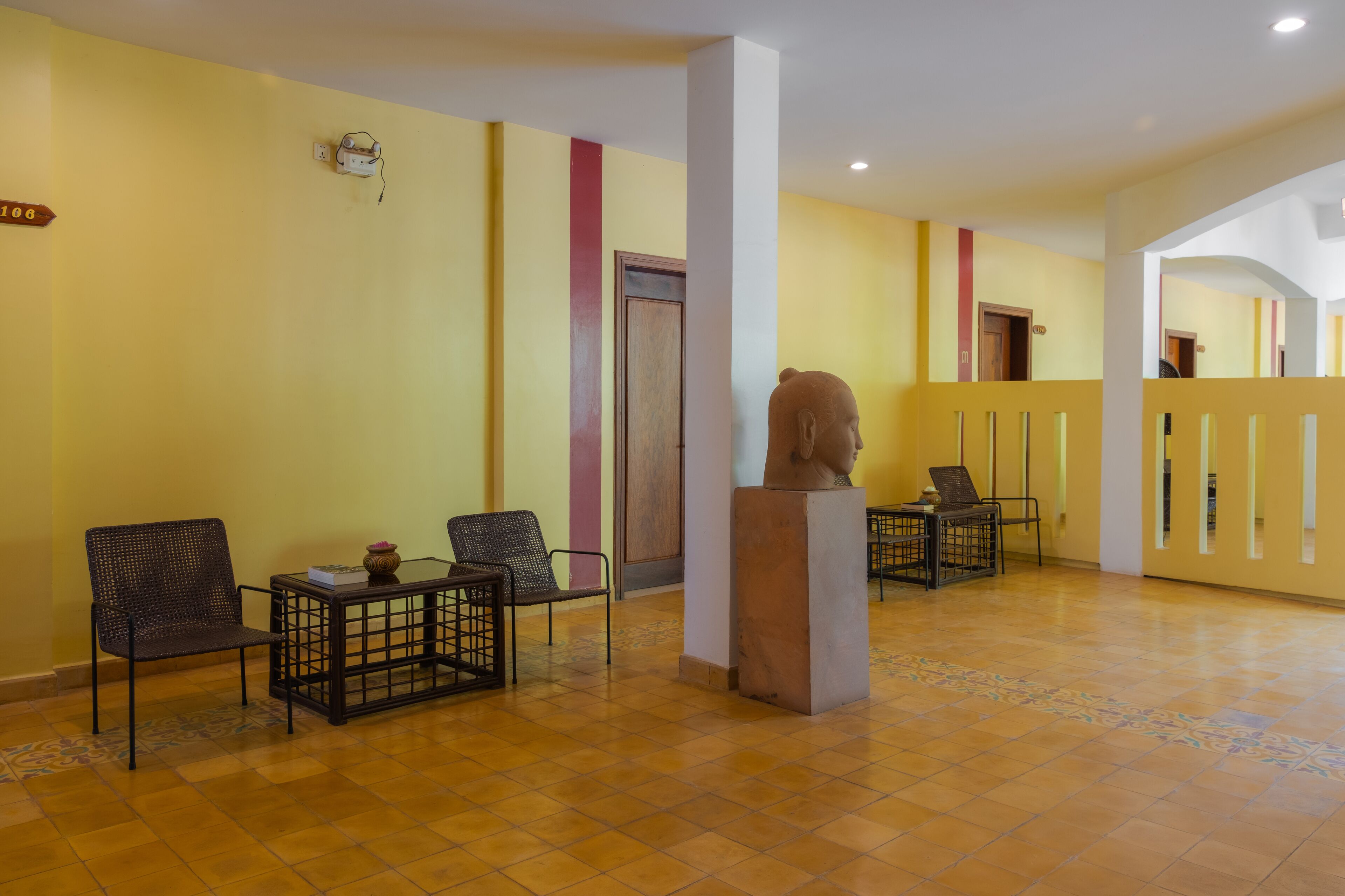 A lobby with yellow walls, a tiled floor, and a bust sculpture. There are two chairs and a small table with a vase.