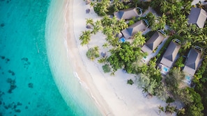 Luchtbeeld van een strandresort met meerdere bungalows, een zandstrand en weelderige palmbomen.