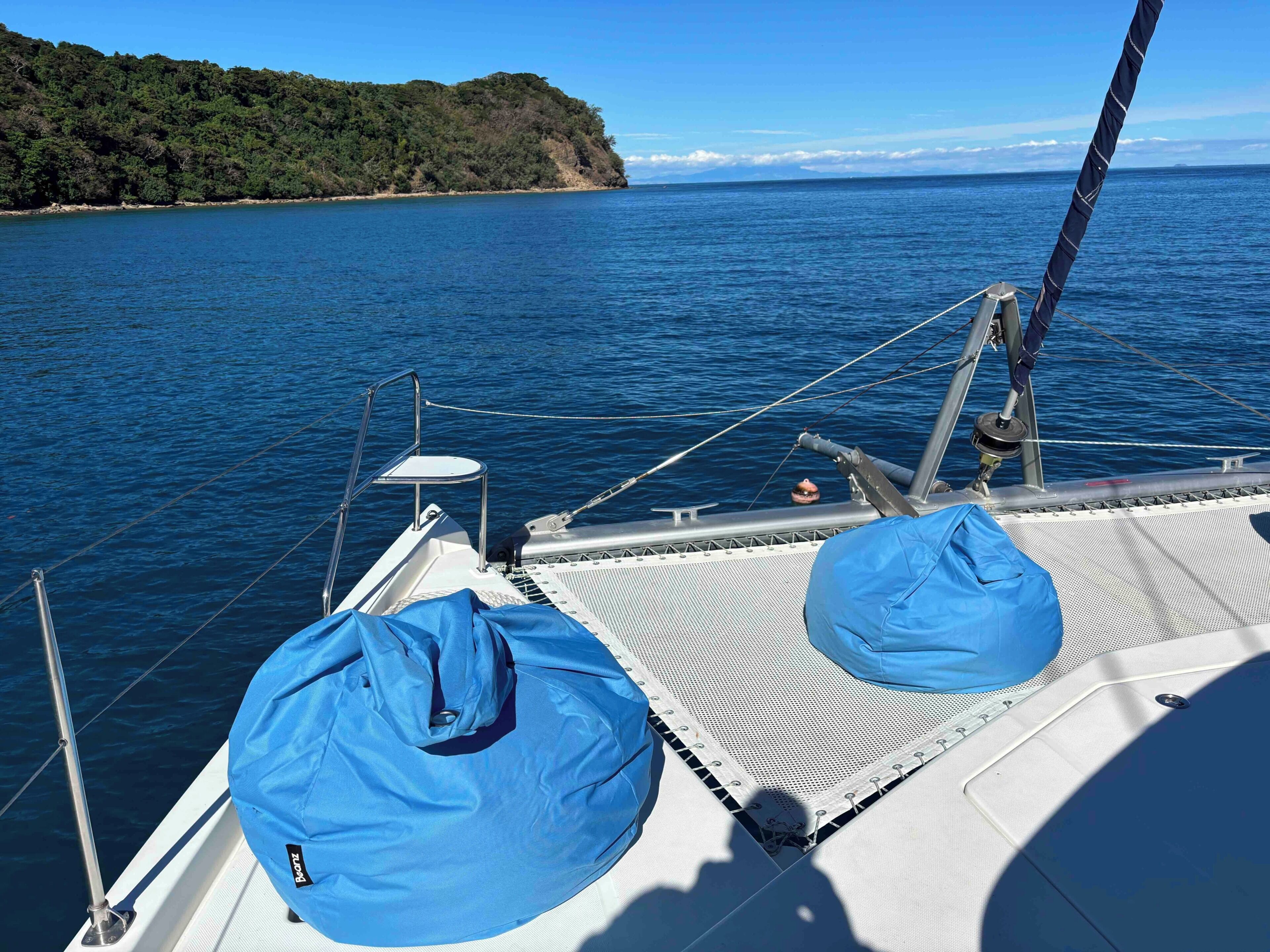 Cubierta de barco con una lona azul, barandales de metal y vista al mar y una isla boscosa.