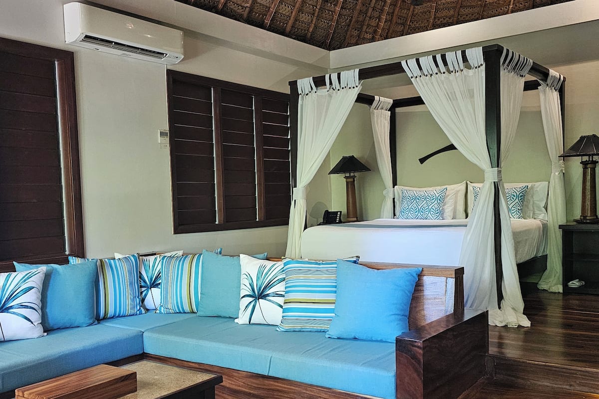 A bedroom with a four-poster bed, a sofa with patterned cushions, a bedside table, and a nightstand with a lamp.