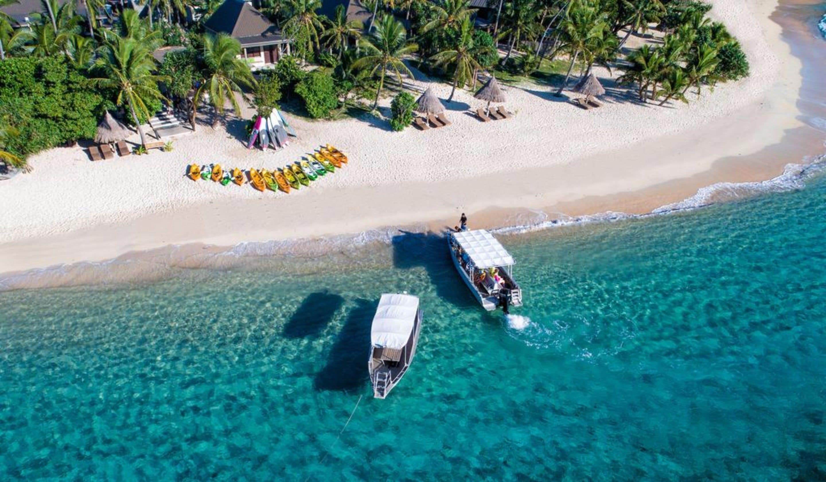 Une vue aérienne d’une plage aux eaux turquoise et limpides, bordée d’une rangée de kayaks colorés, et où l’on aperçoit quelques bateaux ancrés près du rivage.