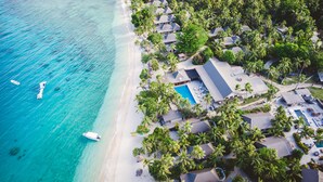 Vue aérienne d’un complexe hôtelier doté d’une plage, d’une piscine et de plusieurs bâtiments de style bungalow, le tout entouré de palmiers.