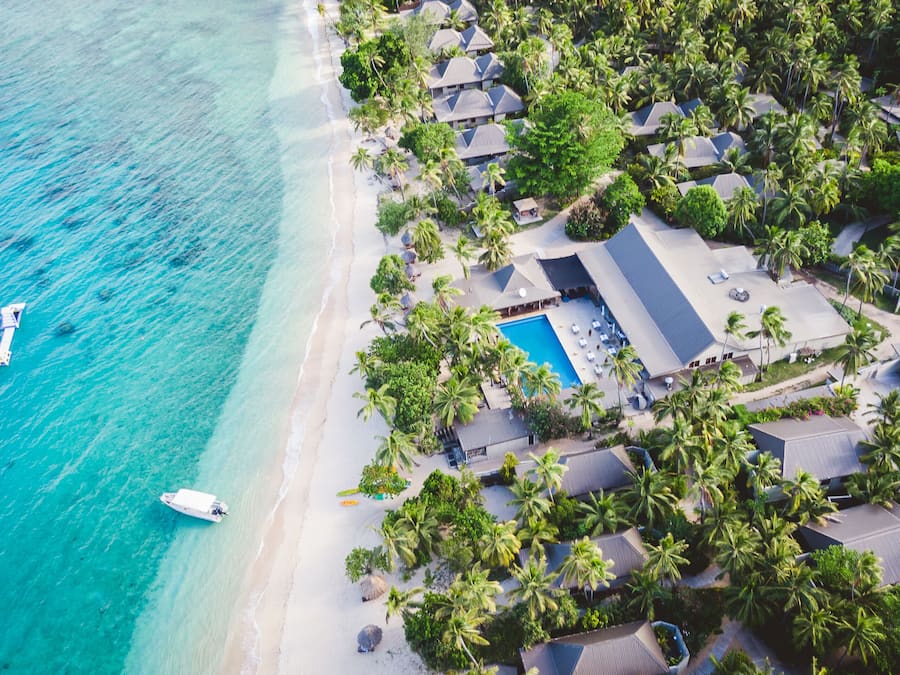 Vista aérea de un complejo turístico con playa, piscina y varias edificaciones tipo bungalow rodeadas de palmeras.