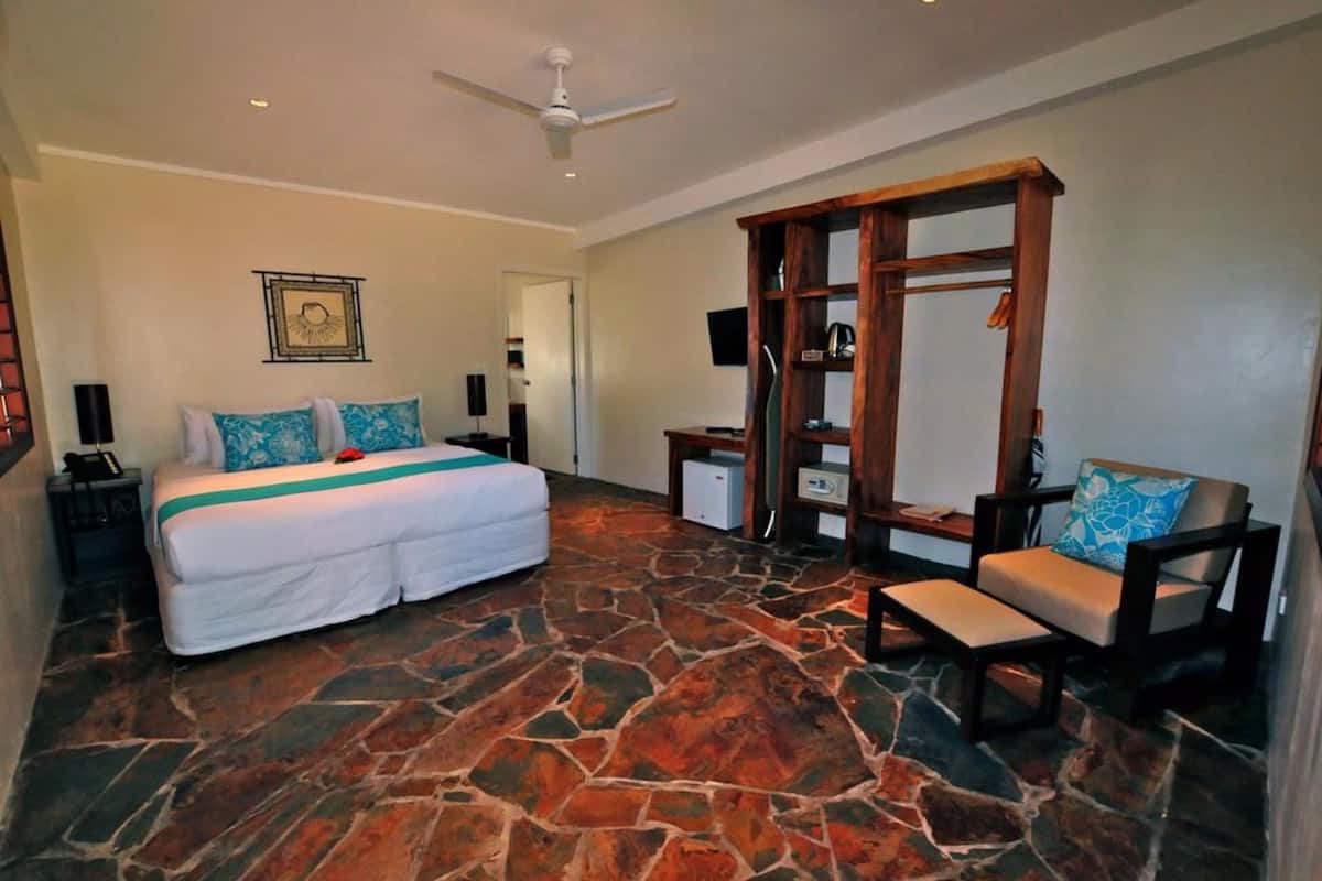 A bedroom with a stone floor, a bed with white bedding, a wooden wardrobe, a chair, and a ceiling fan.