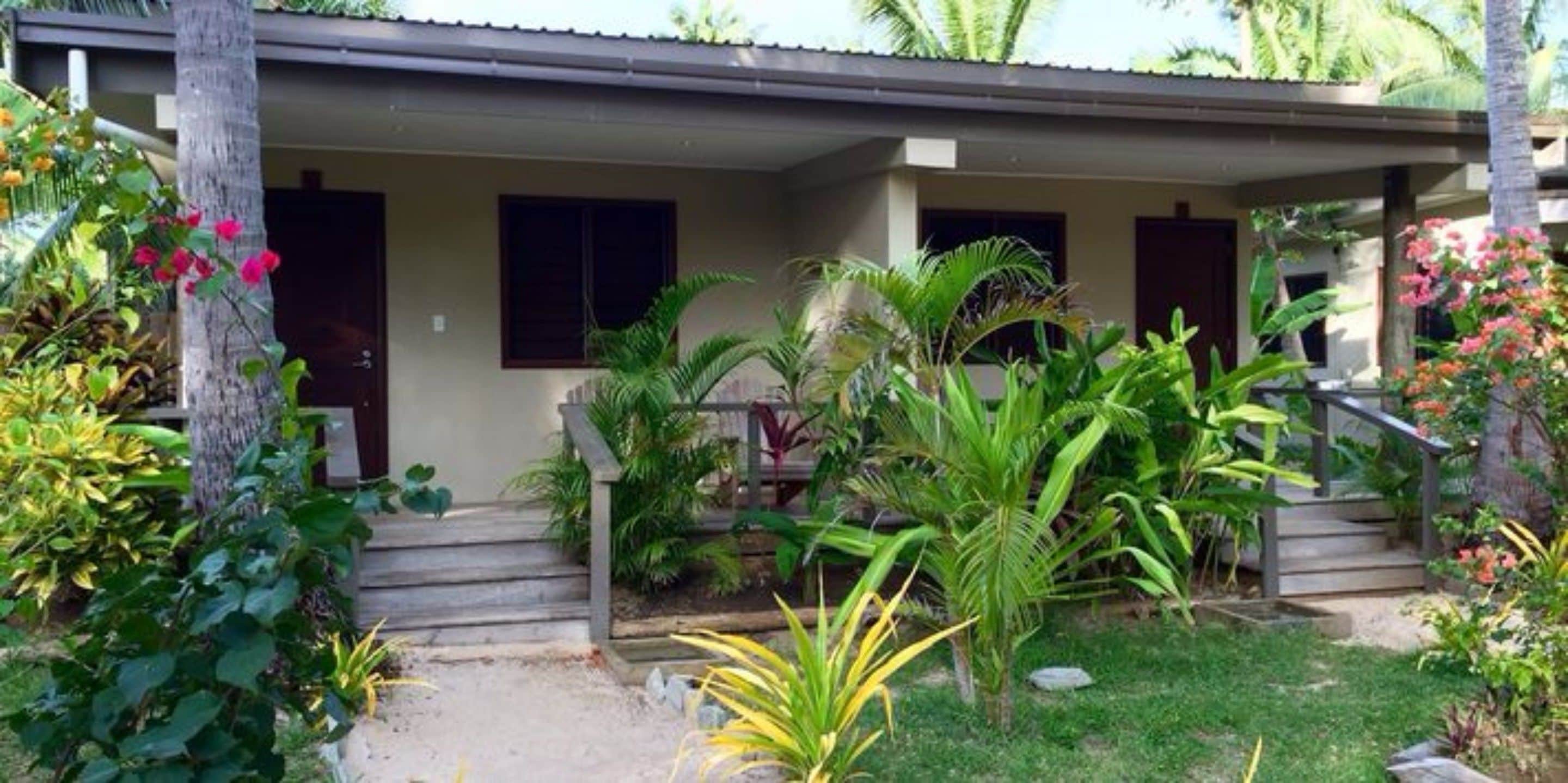 A two-story house with a covered porch, surrounded by tropical plants and a small garden.