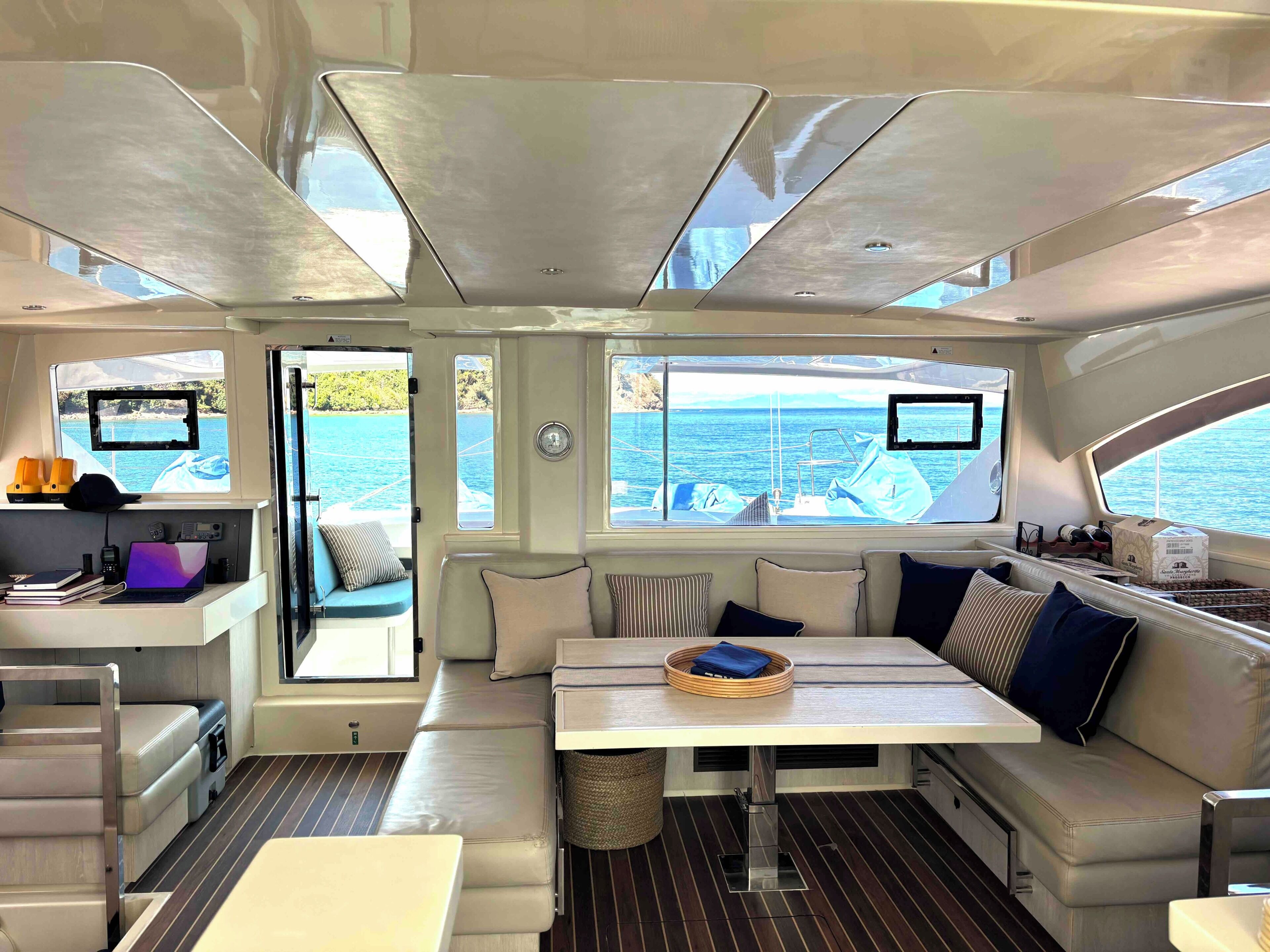 Interior de un yate con comedor, asientos, una mesita y vista al mar.
