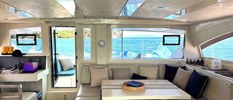 Interior de un yate con comedor, asientos, una mesita y vista al mar.