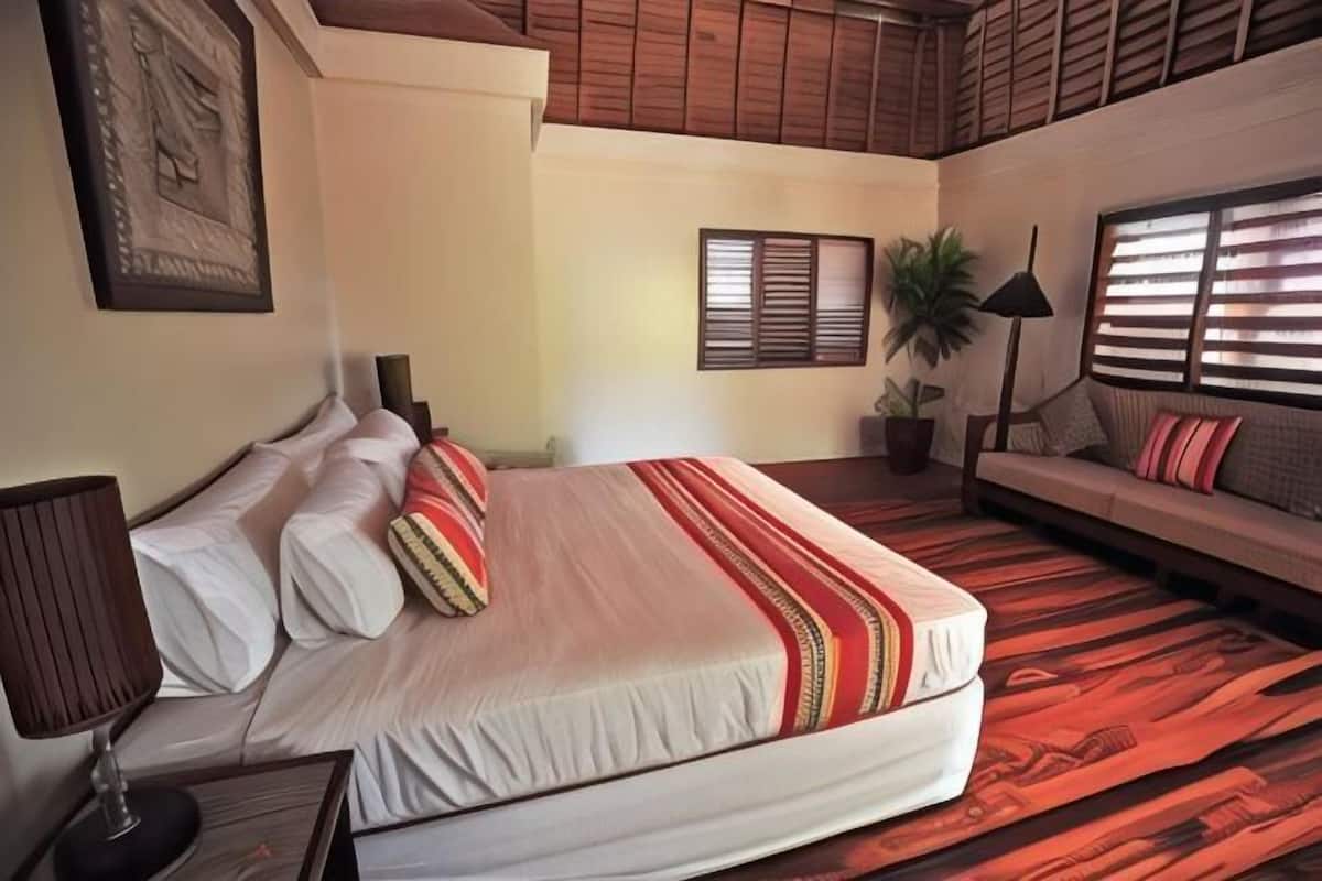 A bedroom with a bed, a sofa, a lamp, a potted plant, and a window with blinds.