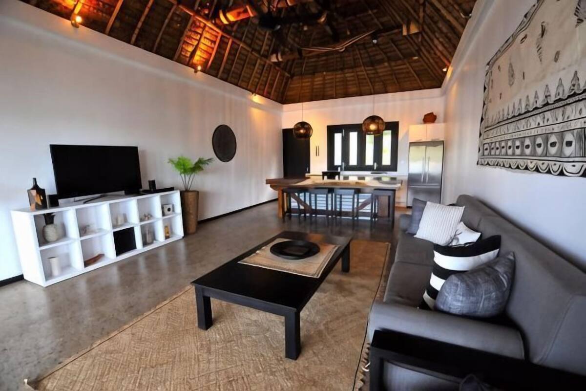A modern living room with a flat-screen TV, a sofa, a coffee table, and a dining area with a kitchen in the background.