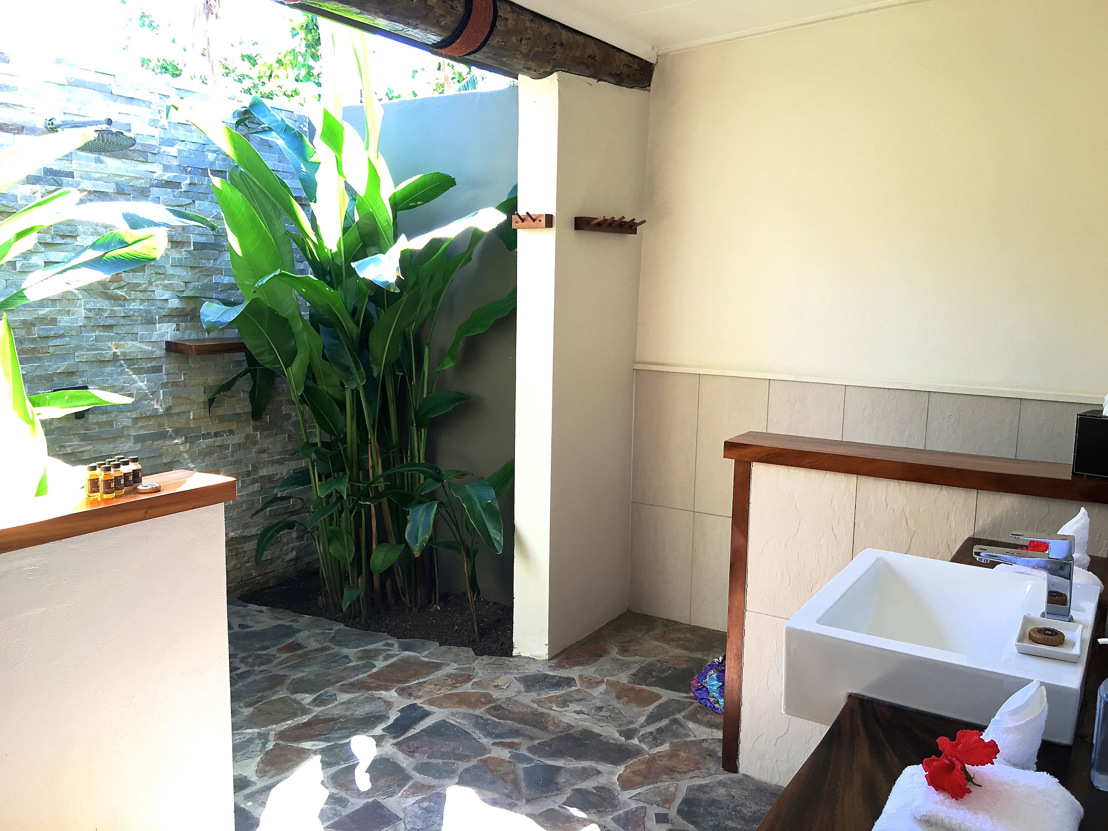 A bathroom with a stone floor, a white sink, and a wooden vanity. There is a large plant near the window and a stone wall in the background.