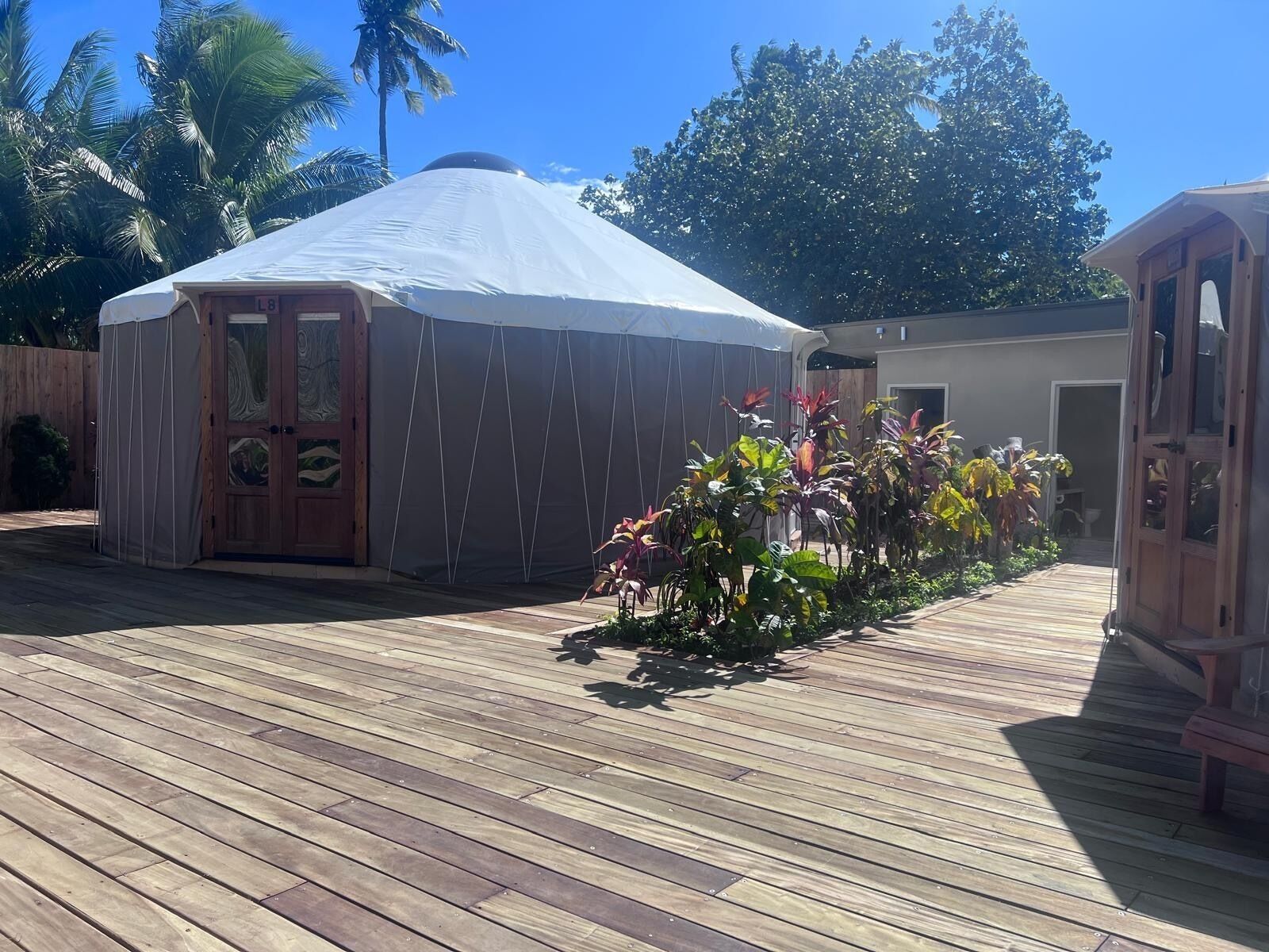 A wooden deck with a covered structure, possibly a gazebo or yurt, surrounded by tropical plants and palm trees.