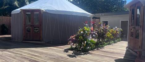 A wooden deck with a covered structure, possibly a gazebo or yurt, surrounded by tropical plants and palm trees.