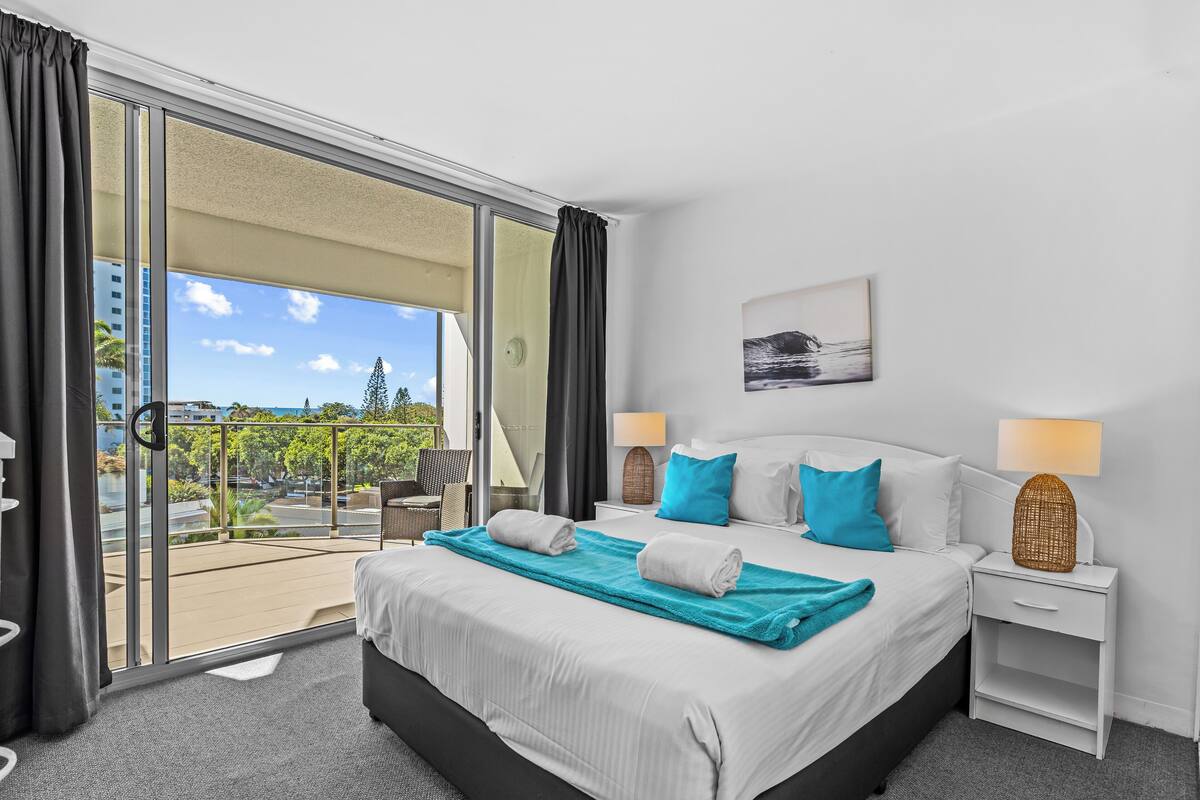 Habitación de hotel con una cama grande, dos mesitas de noche, vistas a un balcón y un cuadro en la pared.