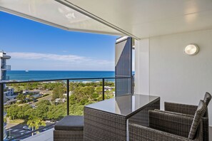 Un balcón con mobiliario de mimbre, una mesa de vidrio y vistas al océano y al paisaje urbano.