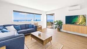 Una sala de estar moderna con un sofá grande, una mesa de centro y un televisor sobre un mueble de madera. Amplias ventanas ofrecen vistas al océano.
