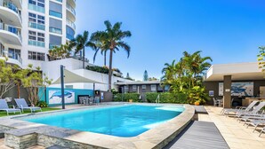 Una piscina con sillas de descanso, palmeras y un edificio moderno con balcones.