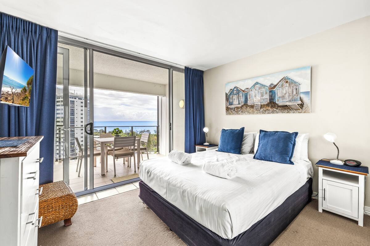 Habitación de hotel con una cama grande, un escritorio, una silla, una mesita y un balcón con vistas al mar y edificios.