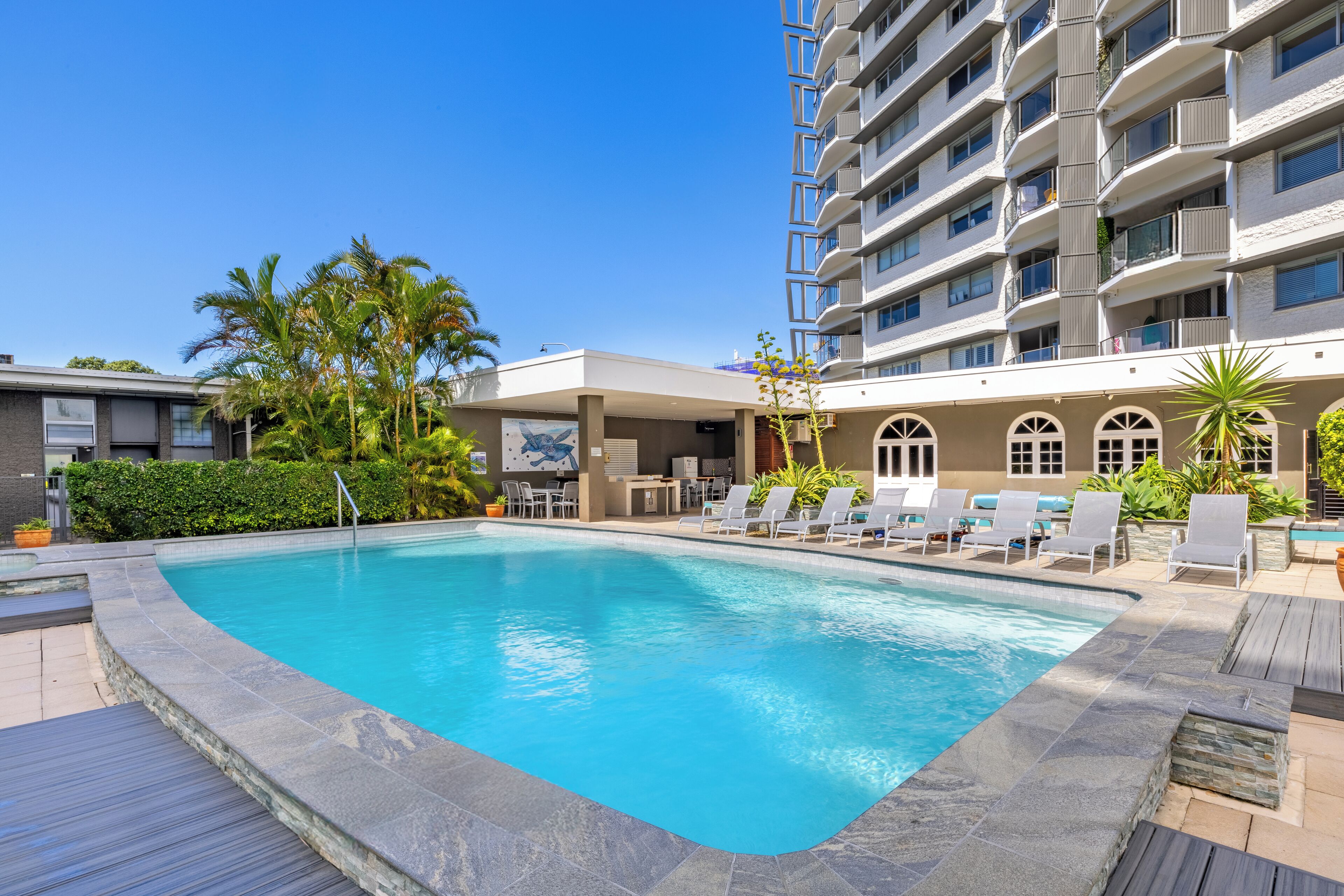 A swimming pool with lounge chairs and a covered area, surrounded by a multi-story building with balconies.