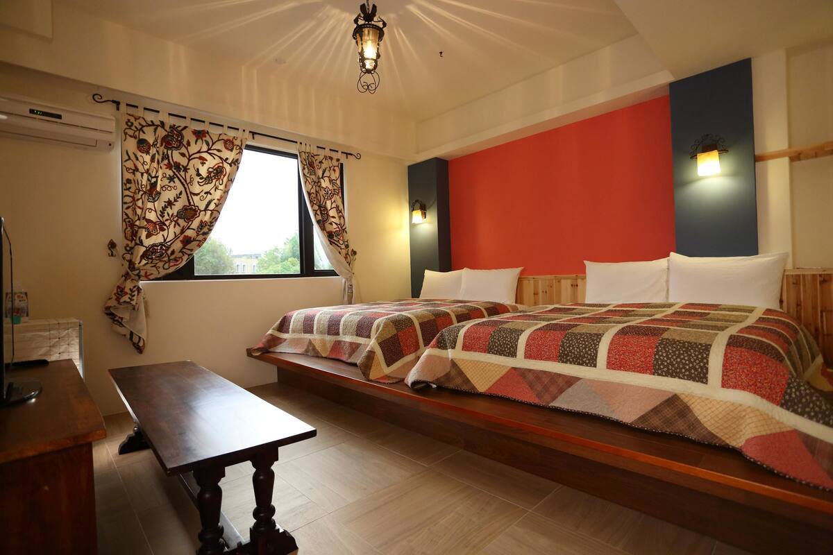 A bedroom with a bed, a wooden bench, a window with curtains, and a wall-mounted light fixture.