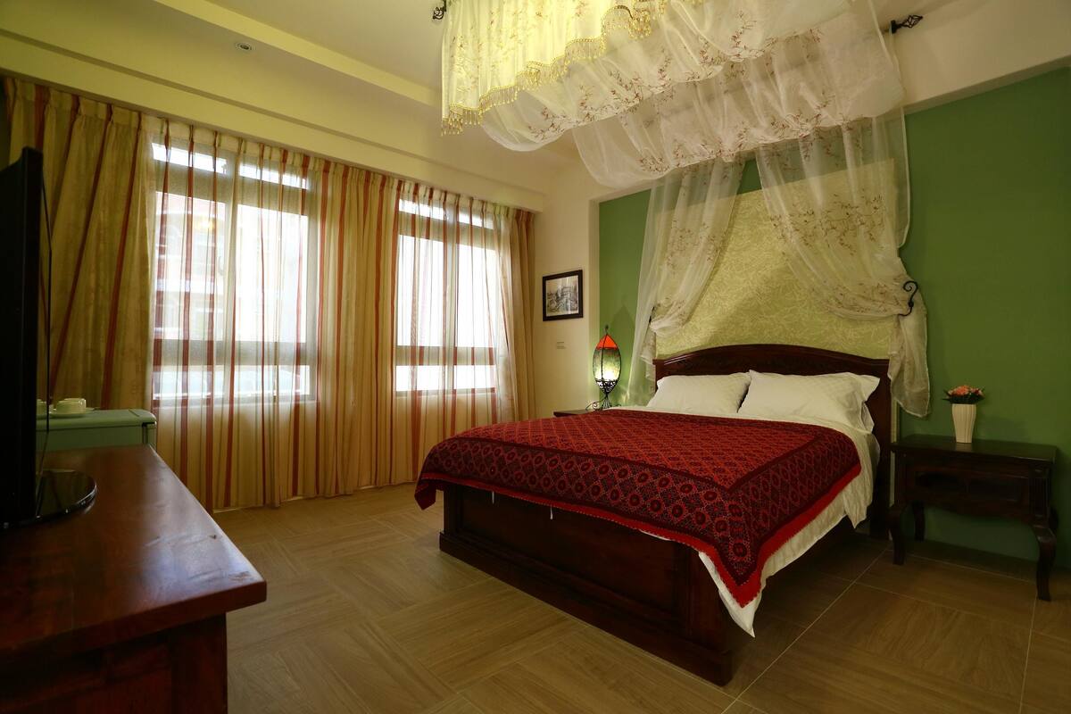 A bedroom with a large bed, a desk, a nightstand, a lamp, and a window with curtains.