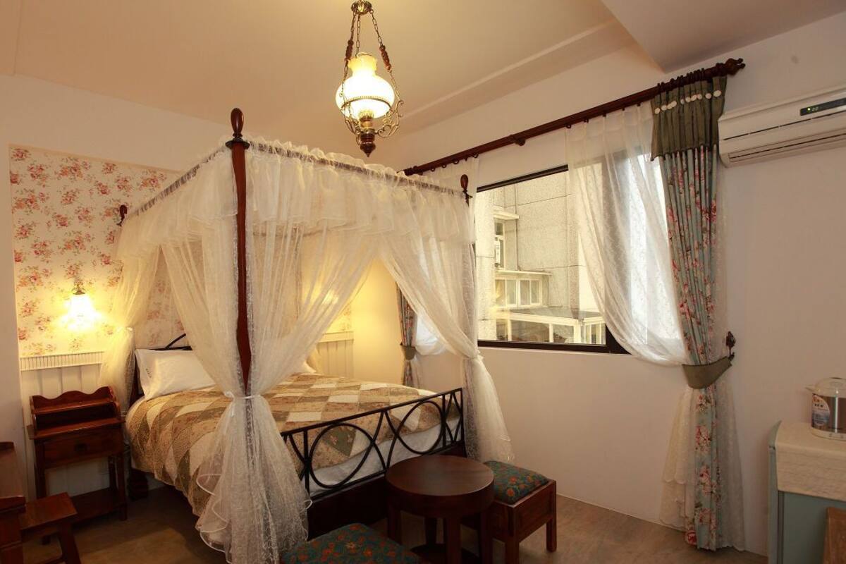 A bedroom with a four-poster bed, a bedside table, a chair, and a small table.