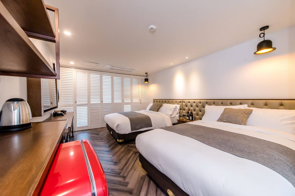 A hotel room with two beds, a wooden desk, a red chair, a kettle, and a minibar.